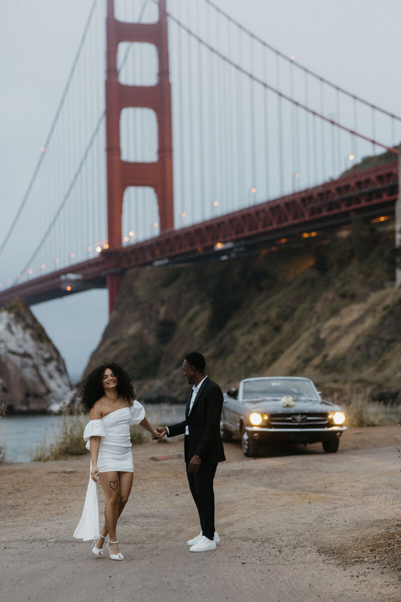 A couple holding hands with a smile, dressed in formal attire, stand in front of the golden gate bridge with a vintage car passing by.
