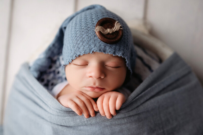 Newborn baby boy tucked into blue fabric with hands peeking out and blue hat on his head