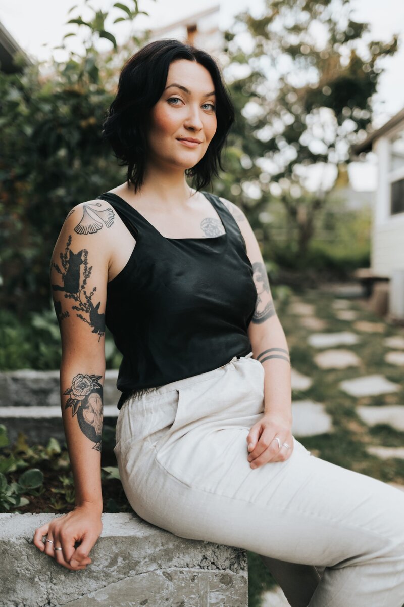 Woman with short, dark hair and several tattoos smirks at camera wearing white pants and a black tank top as she sits on cement wall outdoors.
