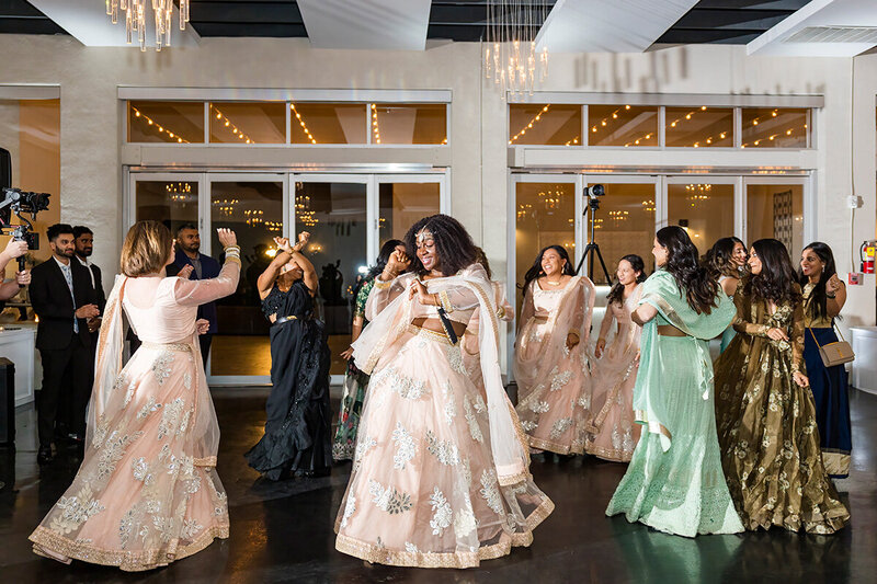 Guests in pastel lehengas dance joyfully on the black-tiled floor at SoHo63, a Chandler wedding and event venue. Surrounded by chandeliers and reflective windows, the celebration brings vibrant energy and cultural beauty to the modern indoor reception space.