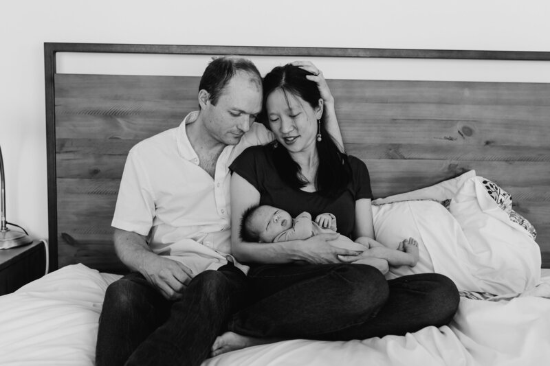 newborn Photographer, a mother and father lean into each other as they admire baby on the bed