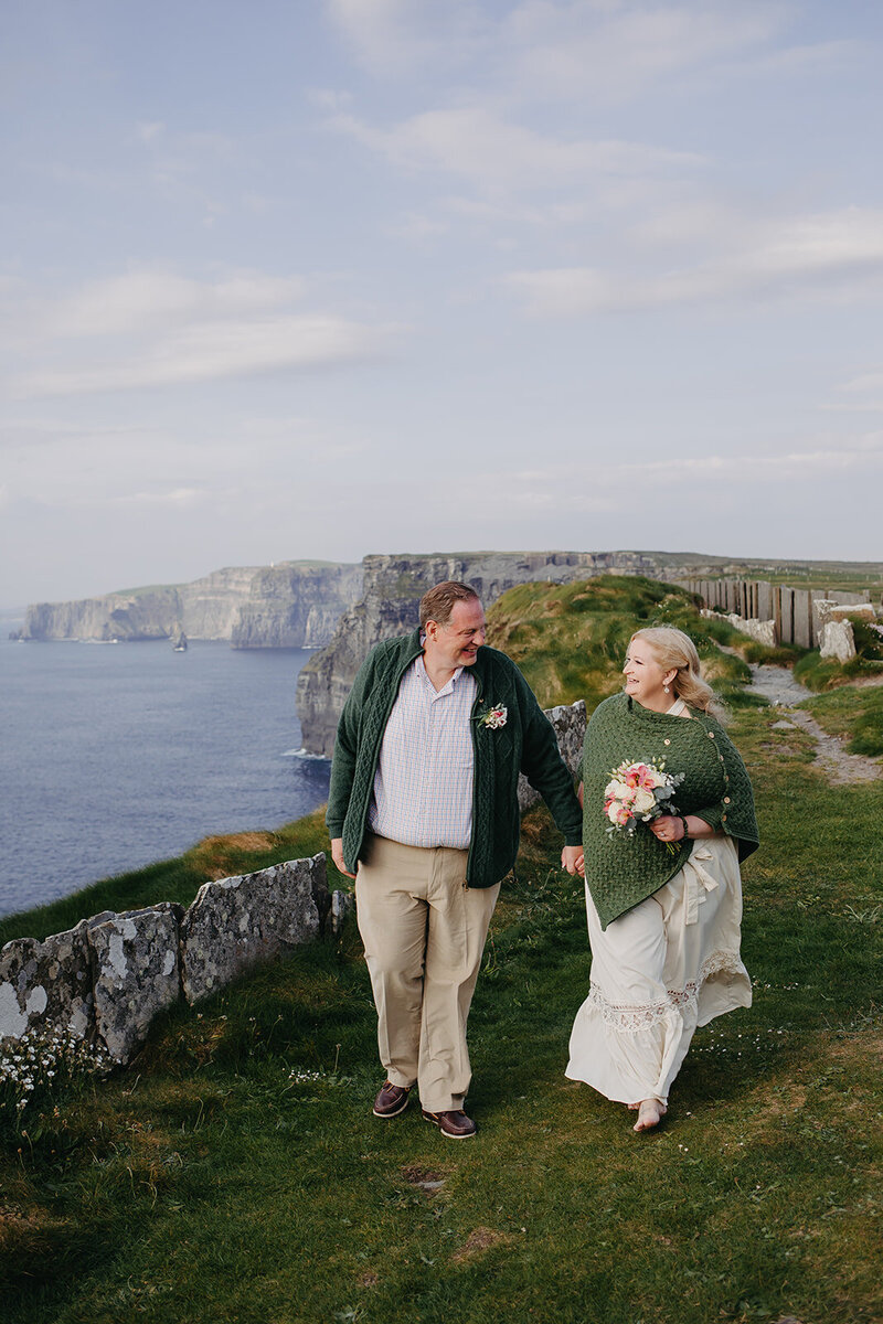 Romantic cliffside elopement of a couple in their second youth, dressed in Irish tones and looking at each other with joy against the stunning cliffs of Moher view. 