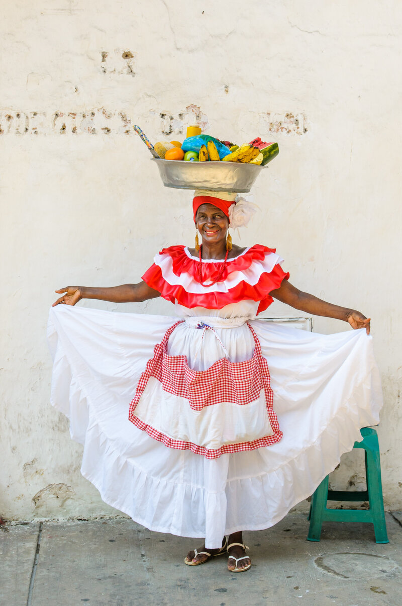 Woman in cultural red and white Columbian  dress stands with bowl of fruit on her head