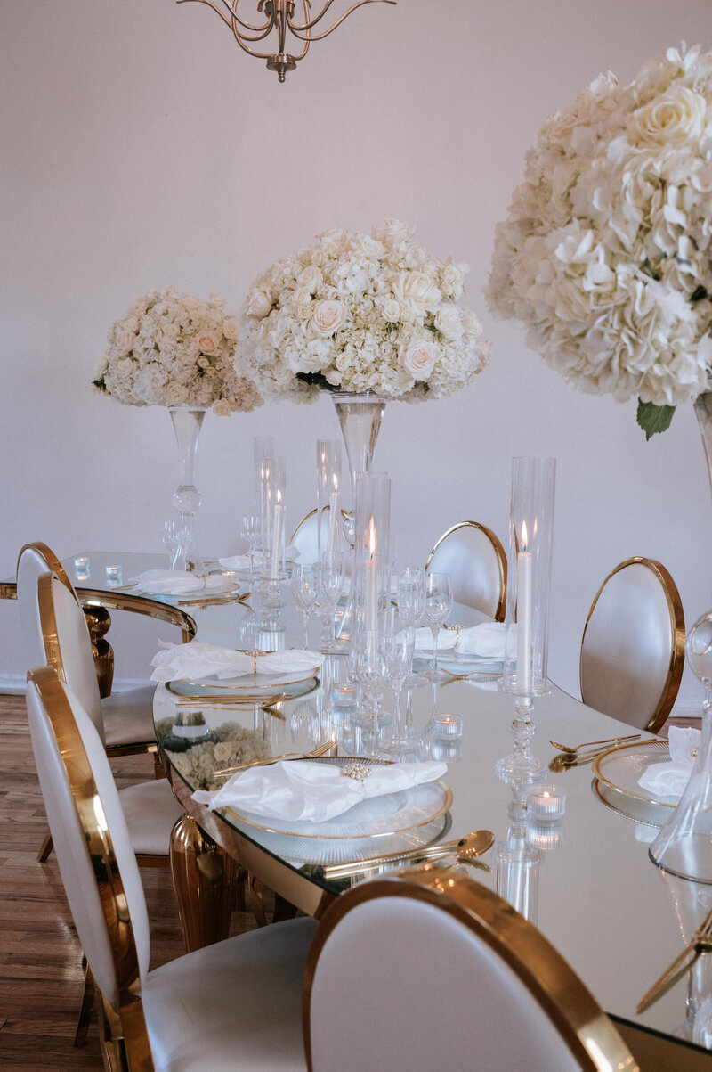 A luxurious wedding reception featuring serpentine. glass-covered tables adorned with white hydrangeas and roses in glass centerpieces, cndlelit sconces, and gold-rimmed Chiavari chairs.   The tablescape is completed with gold flatware and white xharger plates.