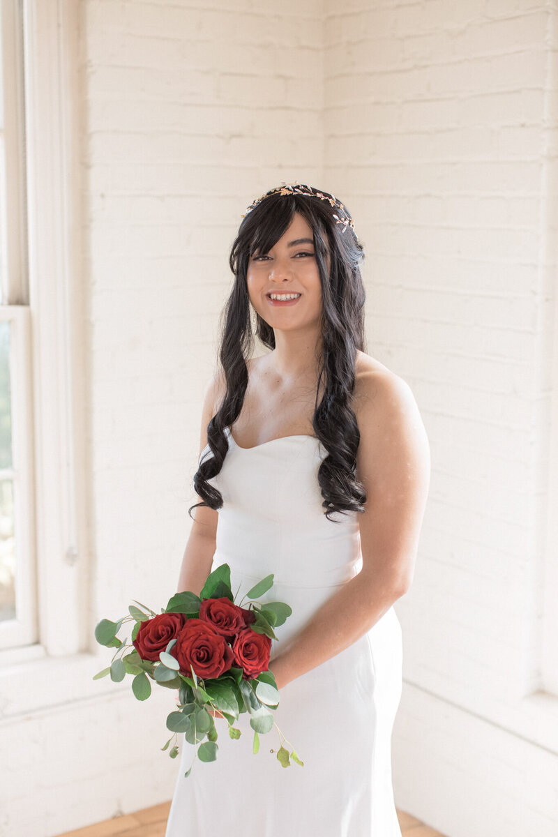 A bride holding red roses and wearing a white dress smiles at the camera during her bridal session 
