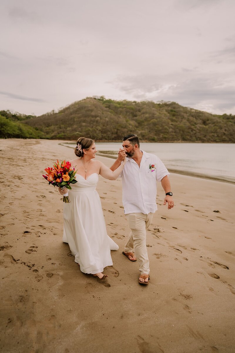 Bride and groom walk hand-in-hand along a sandy beach in a tropical destination, with lush green hills in the background.