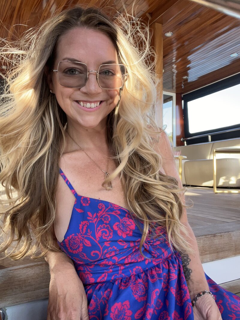 Marcie Muensterman is holding a glass of red wine. She has glasses on and long blonde hair. She is wearing a gray wrap over a dark top. She is leaning against a deck railing.
