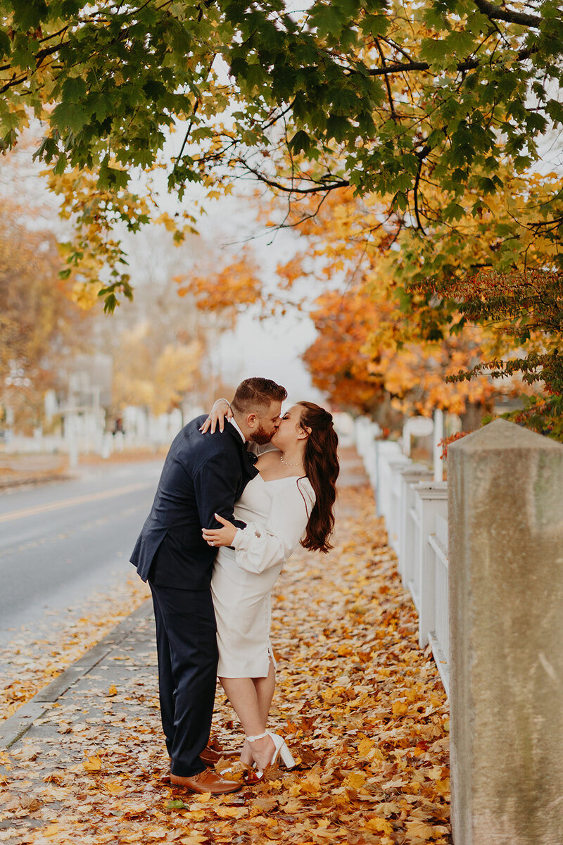 Autumn city elopement: The bride and groom share a kiss on a road adorned with fallen leaves, creating a picturesque scene.