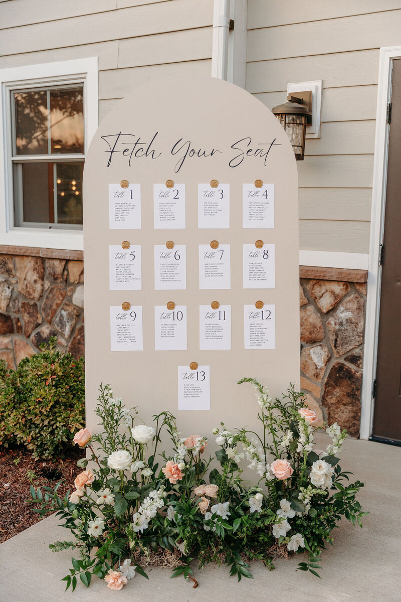 A wedding seating chart stands against a wall, titled "Fetch Your Seat." It features lists from table 1 to 13 with guest names. The base is adorned with lush white and peach flowers and greenery, perfectly set for picturesque Leesburg VA wedding venue images.