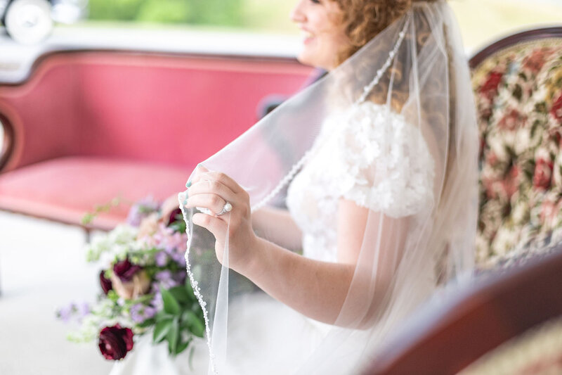 A bride adjusting her veil while seated on a vintage floral sofa, surrounded by soft natural light and floral arrangements, at Willow and Stone Events near Charlotte, NC.