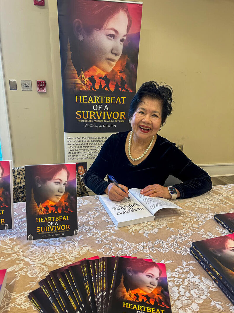 Author and speaker Nita Tin at the Scenic City Women’s Network Lydia Award event in Chattanooga, Tennessee, smiling as she signs copies of her book Heartbeat of a Survivor. She is seated at a table decorated with a lace-patterned tablecloth, surrounded by displayed copies of the book, with a banner featuring the cover image in the background. She wears a black dress with pearls and holds a pen in hand.