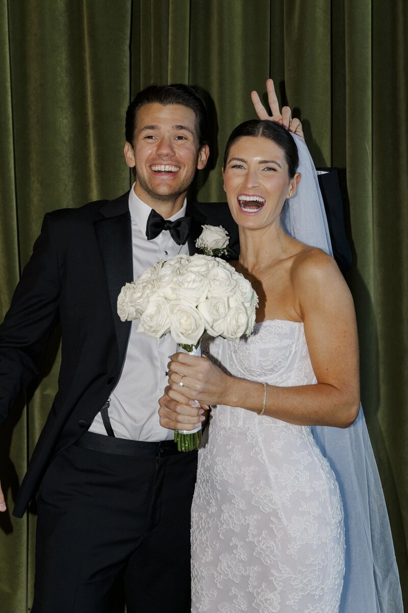 Bride and groom laughing together in front of a green velvet curtain with a white bouquet