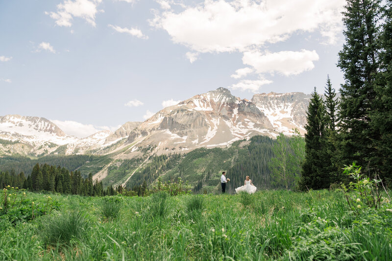 A man holds a woman in a wedding dress in front of a lake and mountains