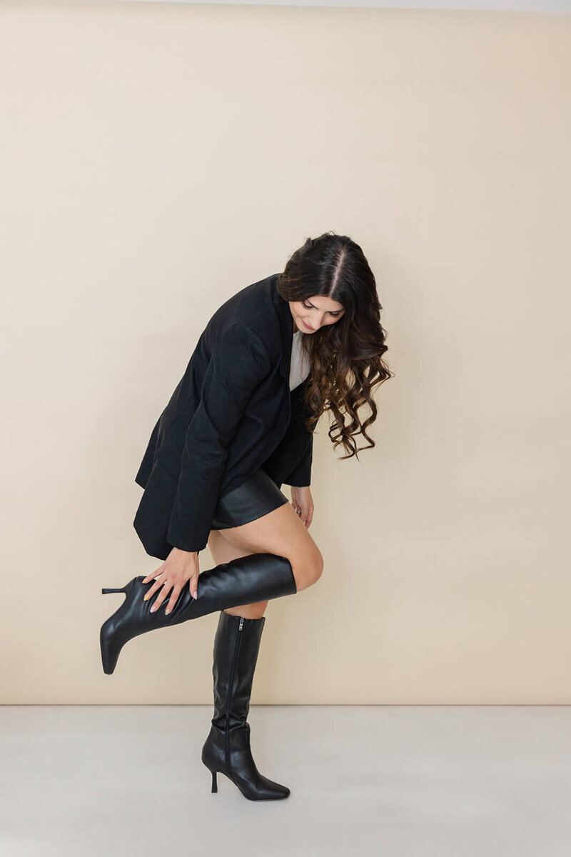 Bel, the founder and owner of Soulve Studio, is adjusting her knee-high black boots while dressed in a chic black blazer and skirt. Her long wavy hair cascades over her shoulders as she strikes a stylish pose against a minimalist beige background, reflecting her modern and confident personal style.