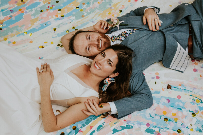 Bride and groom laying on hand-painted rainbow color backdrop with confetti in Atlanta GA backyard wedding