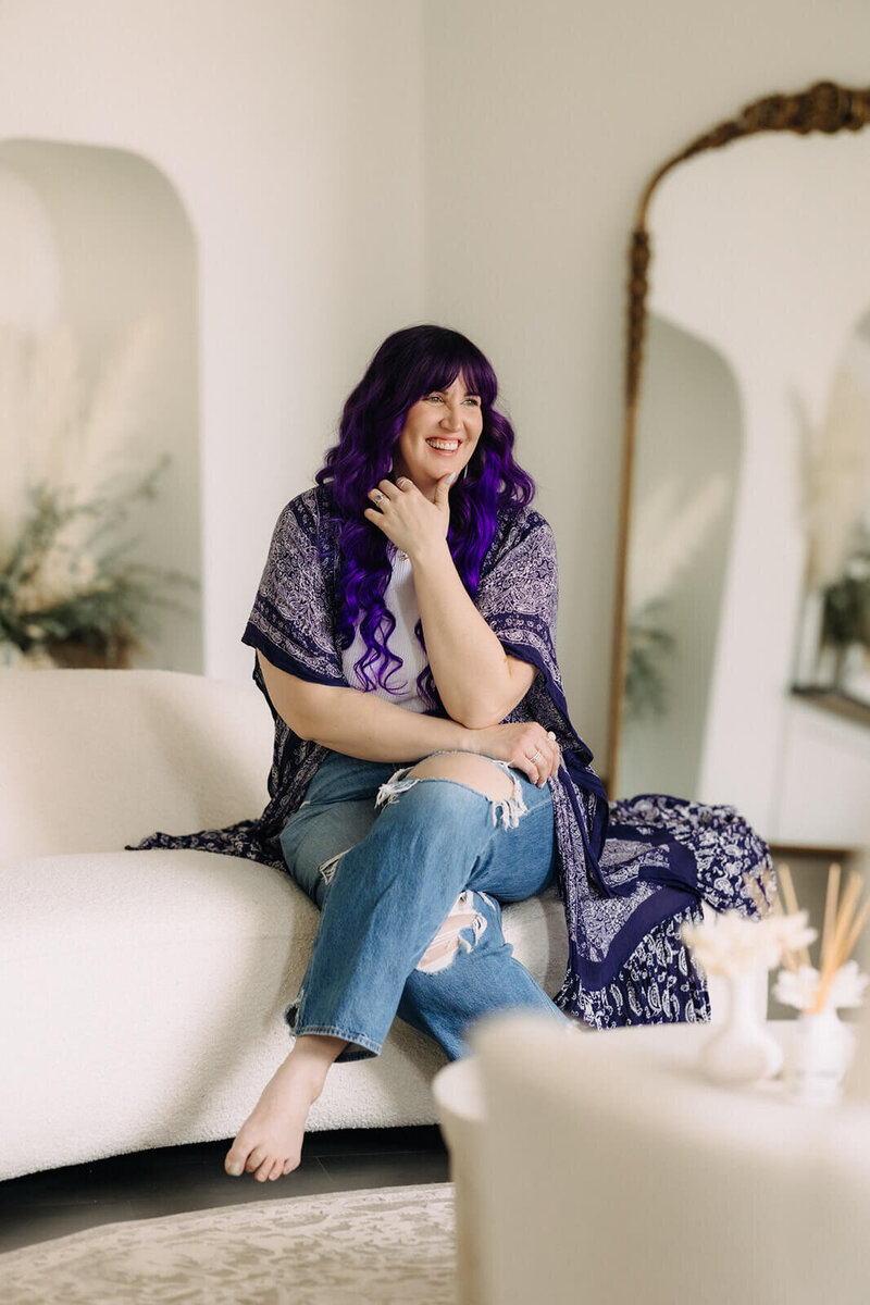 Sarah sitting on white sofa, with purple hair , ripped jeans and purple wrap. Finger Lakes wedding photographer, New York