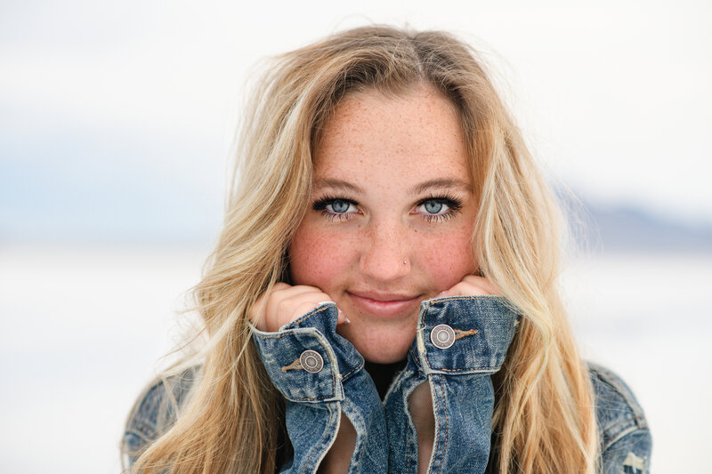 A Denver senior girl with bright blue eyes and her hands up by her face wearing a jean jacket looking intensely at a Denver senior photographer