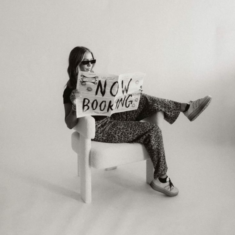 Woman holding a “Now Booking” newspaper—symbolizing open coaching spots at The Clair Agency for women ready to shift their dating story.