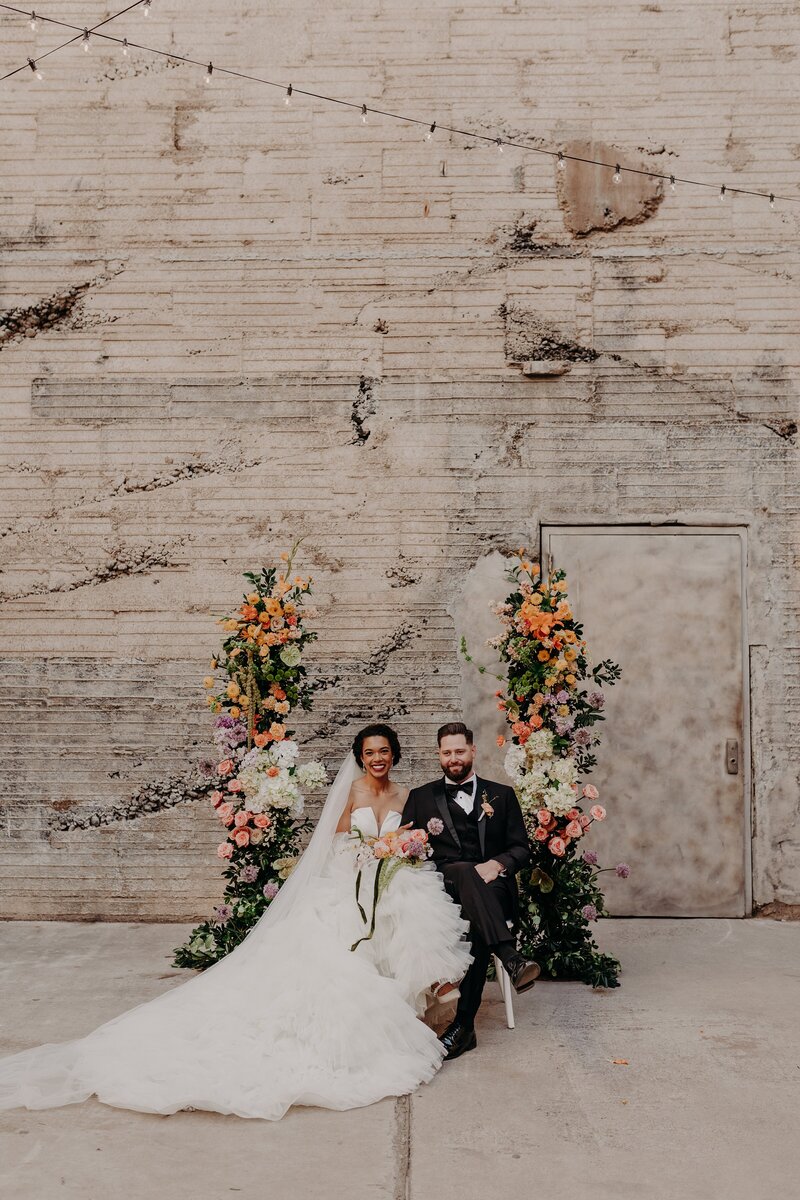 Bride and groom sit side by side in-between floral arch