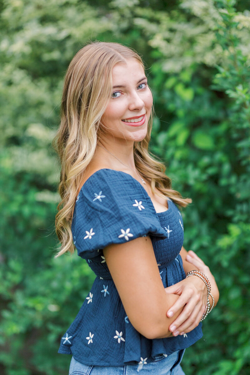 Senior portrait of a smiling teen girl in a navy floral top, surrounded by lush greenery, taken by Raleigh Senior Photographer Lindsey Lambert Photography.