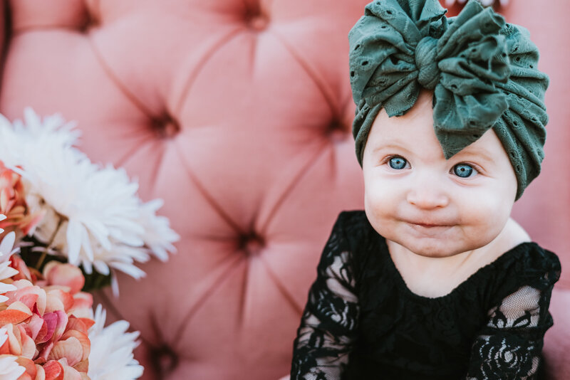 Baby girl in black and green bow sitting in pink chair for milestone photos.