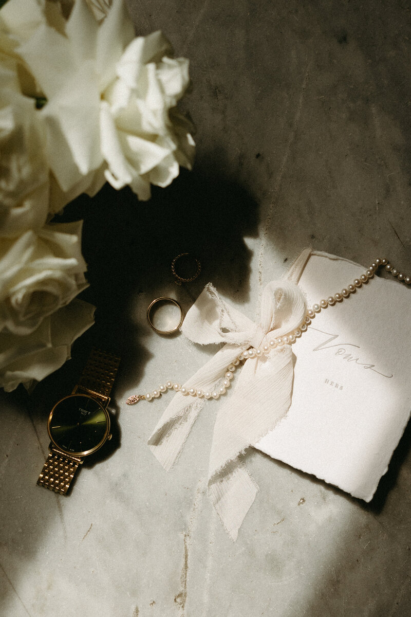 Flat lay of bride and groom's rings, white roses, vow books and pearl necklace. 
