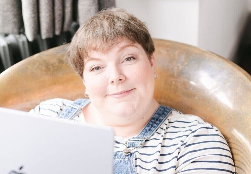 Lucy from Disobedient Business® Co. smiles warmly while reclining in a copper chair, wearing a striped top with denim dungarees and working on a laptop in a bright, cosy space.