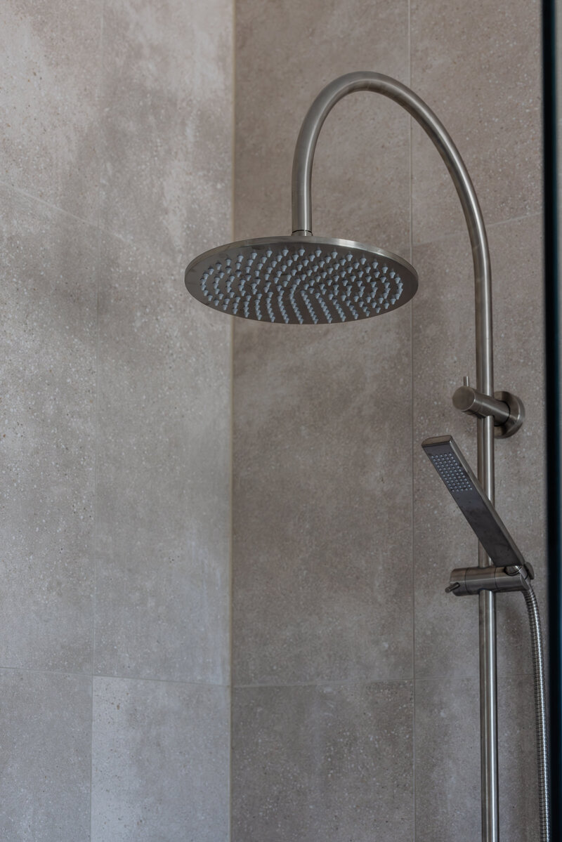 A sleek, modern showerhead with a curved metal arm mounted on a light stone wall, crafted with precision by Newcastle Kitchen and Bathroom Designer.