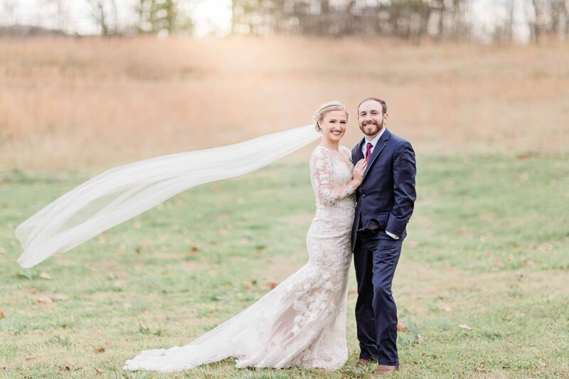 Bride and groom in a field with her veil blowing in the wind.