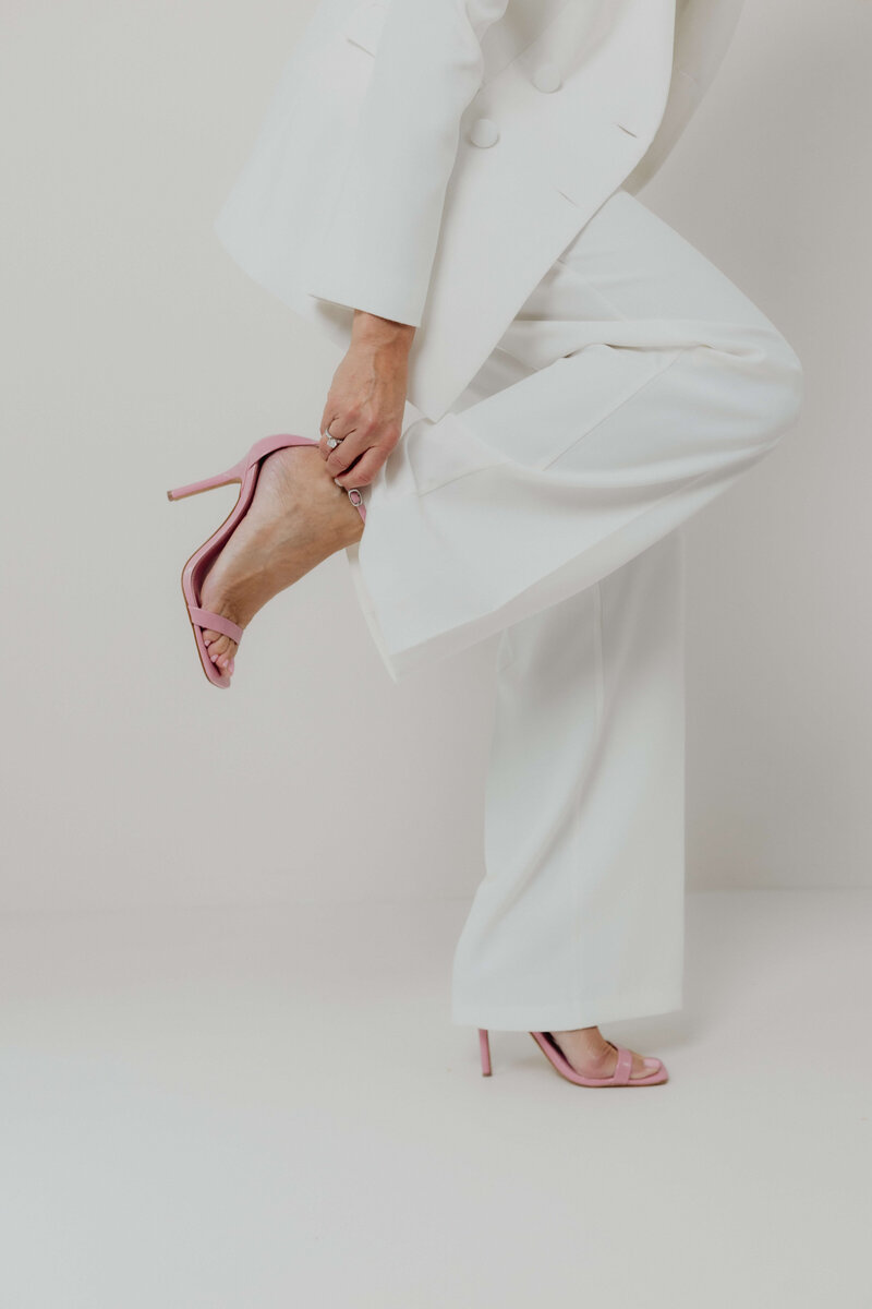A close up of a women in a white power suit and pink heels with her leg up .