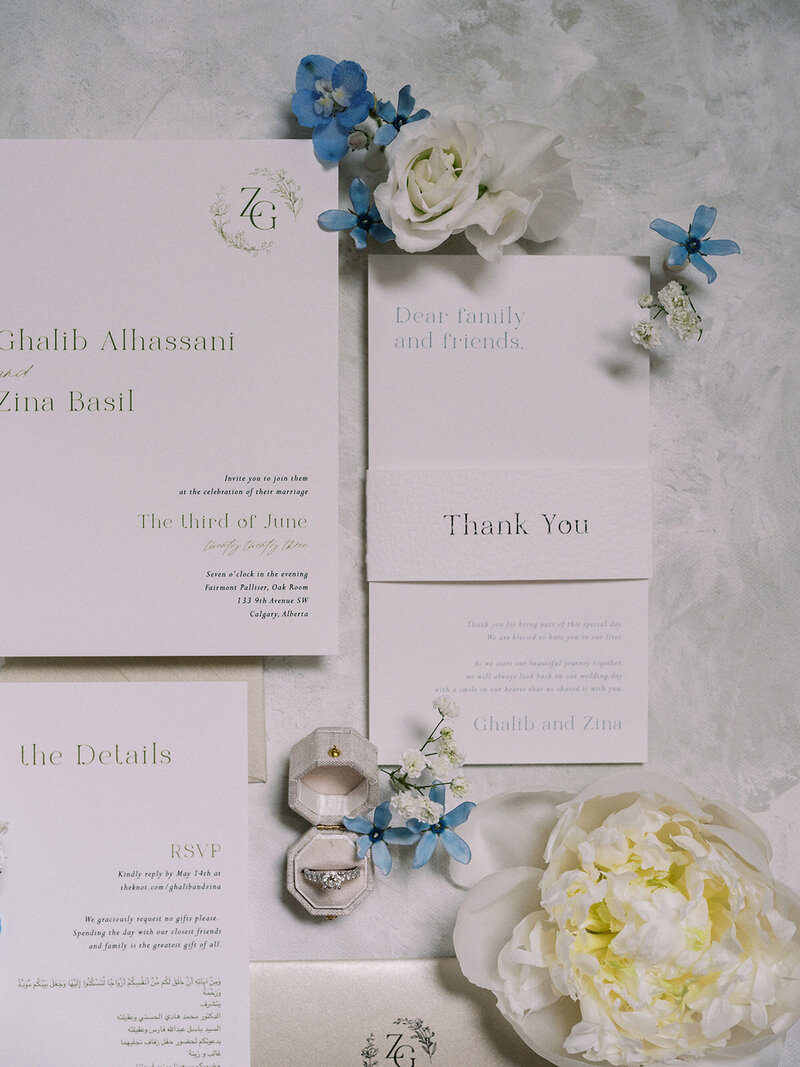 Wedding invitation set for Ghalib Alhassani and Zina Basil, designed by a full service wedding planner. Includes an invitation card, thank you note, details card, RSVP card, and rings in a ring box, all accented with white and blue flowers.