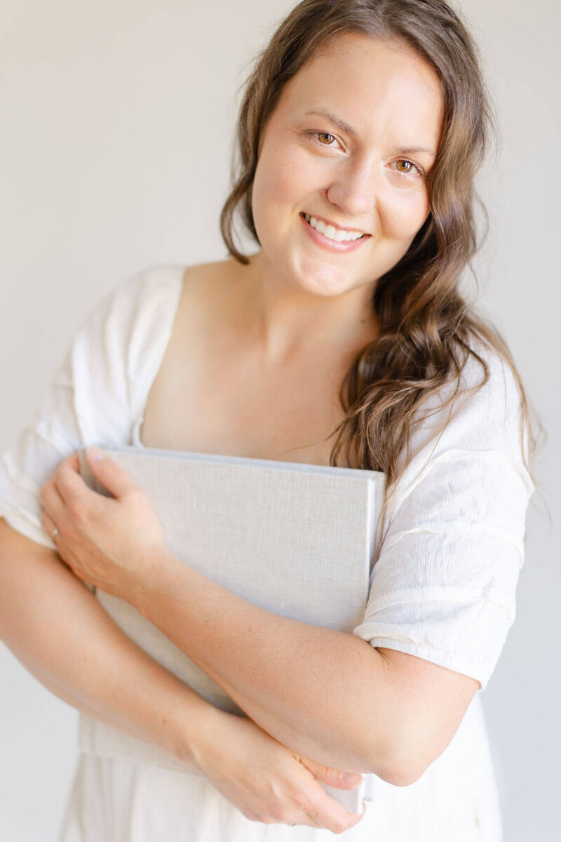 Woman with long brown wavy hair smiling at the camera and holding an heirloom linen album close to her chest.