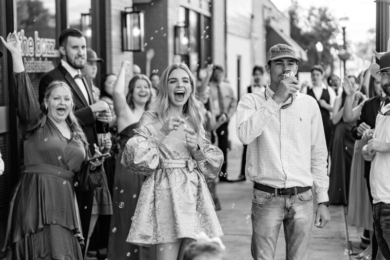 A group of people cheer and blow bubbles outside a building. A smiling woman in a light patterned dress and a man in a cap stand at the center, surrounded by joyful onlookers. The scene appears festive and celebratory.
