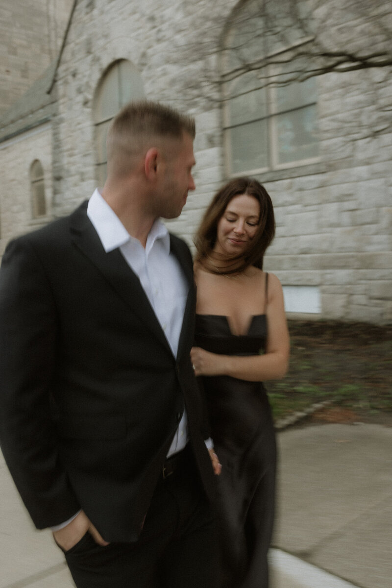 A couple walking past a stone building, the woman in a black dress smiling at her partner during an intimate moment.