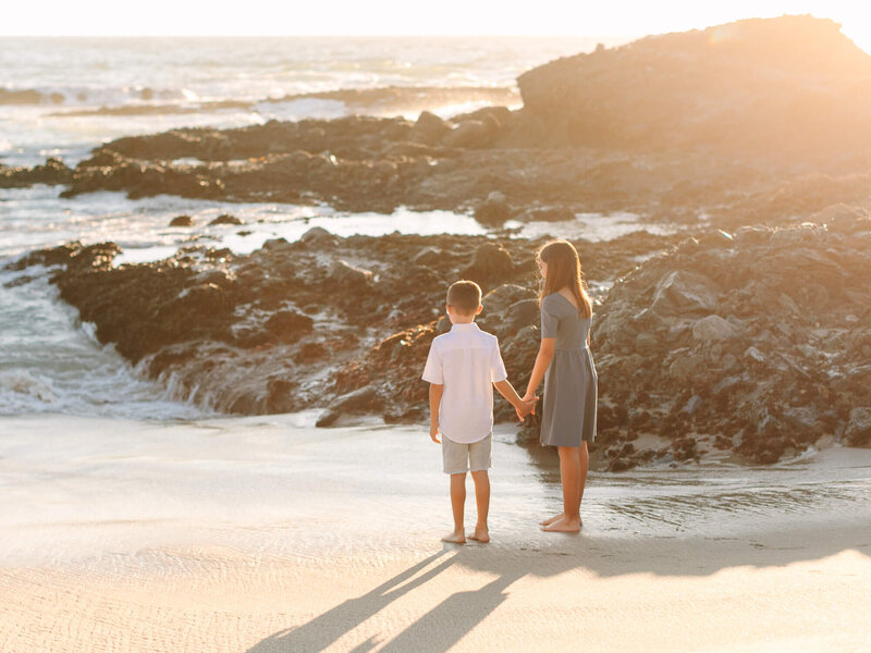 Two children holding hands, standing barefoot on a sandy beach at sunset. The ocean waves and rocky shore create a serene, tranquil scene.