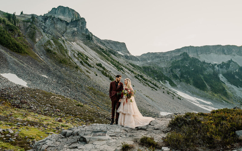 a couple in their wedding attire gaze lovingly at each other as they stand in a beautiful mountain landscape 