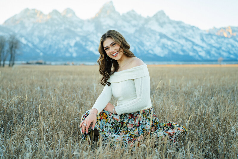 Senior portraits in Grand Teton National Park. A girl sitting in a grass field with one hand on her knee and the other hand on her thigh. Her head is slightly tilted and she's smiling at the camera. Behind her are the Grand Tetons.
