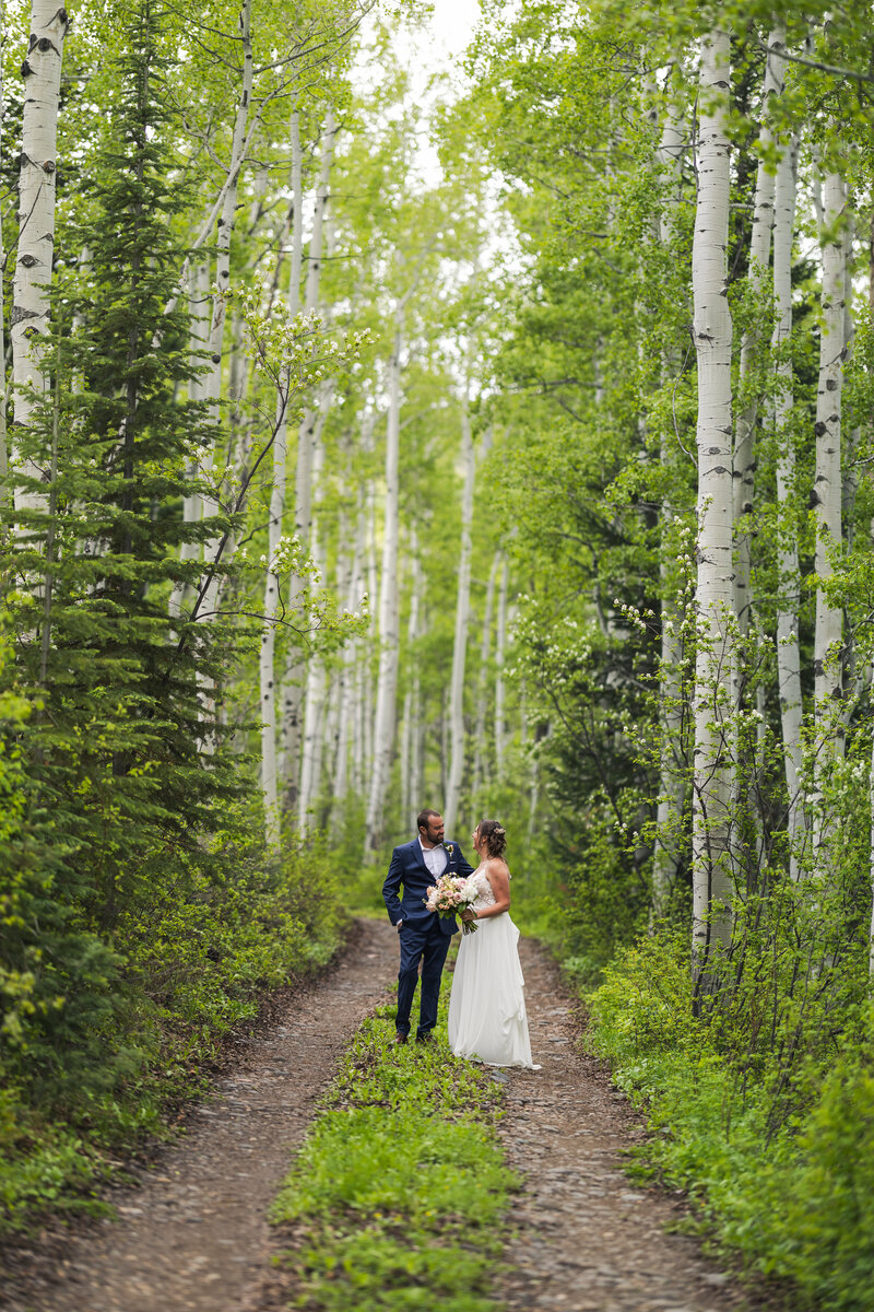 A bride and groom walking on a road through a thick aspen grove