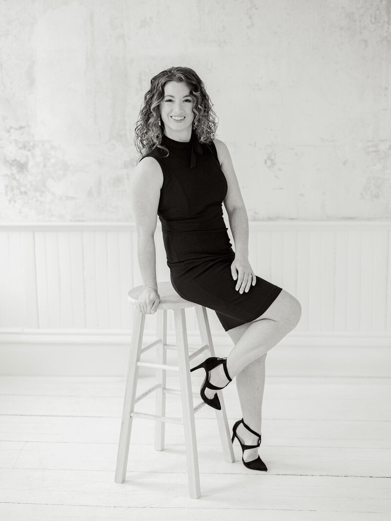 Black and white headshot of Lauren Baker in a black dress and heels sitting on the edge of a stool