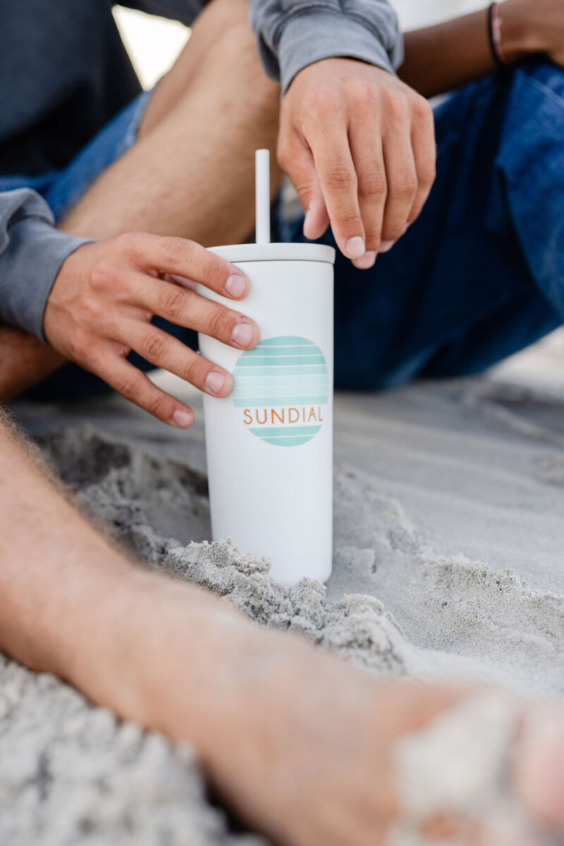 a custom designed white sundial tumbler with a straw sitting in the sand