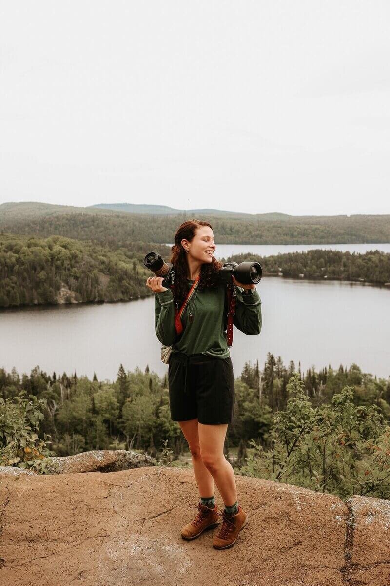 The photographer standing on a rock in front of a lake holding her cameras