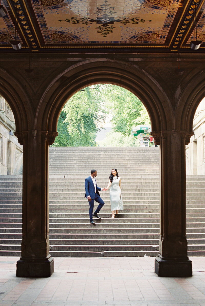 Engaged couple walking hand-in-hand on the iconic steps of Bethesda Terrace in New York City, framed by ornate arches and vibrant greenery, captured by Stephanie Michelle Photography, a luxury wedding and engagement photographer.
