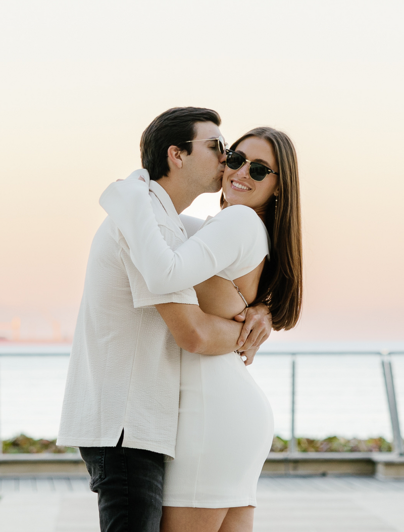 Happy engaged couple in sunglasses, embracing and kissing at sunset in Miami, Florida, captured by Claudia Amalia. Claudia is a wedding and lifestyle photographer based in Miami and Florida Keys, South Florida. She offers stunning engagement packages and specializes in destination weddings, providing beautiful and memorable photography services.