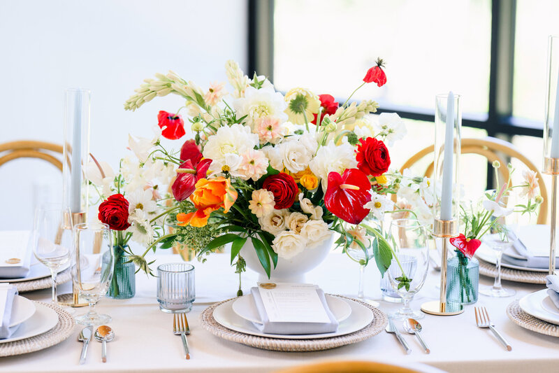 Bright floral centerpiece with pops of red, orange, and cream on a wedding reception table, designed by a Dripping Springs wedding planner.