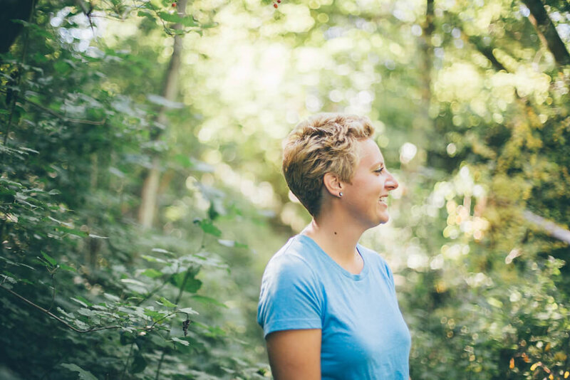 Writer, Sarah Freymuth, stands among trees in a blue t-shirt smiling and looking into the distance