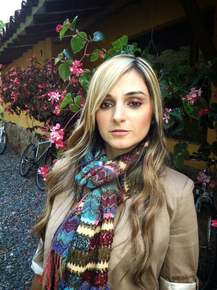 Image of Juliana Soto, the owner of Sinful Sweets Cakery. Juliana is wearing a scarf and tan blazer and standing in front of flowers outside.