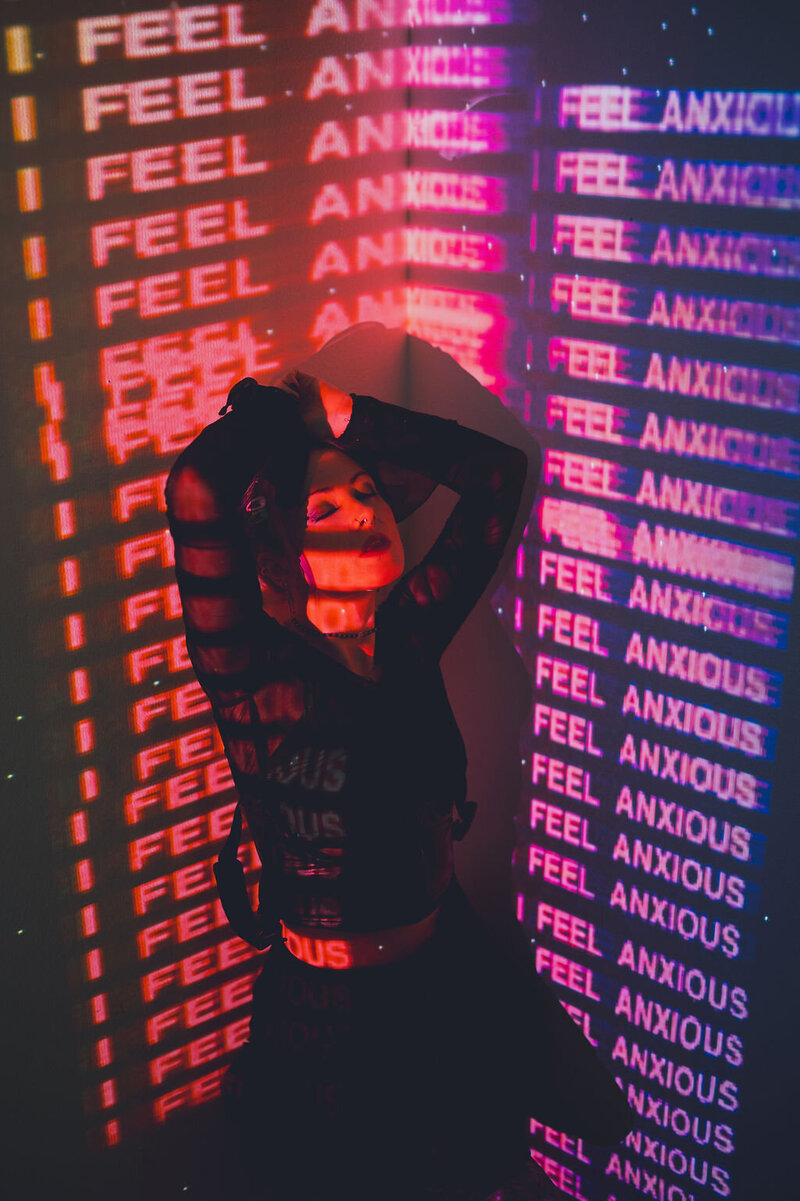 Woman stands with arms overhead as she closes her eyes. Pink, purple, and red projected light reads "I feel anxious" repeatedly.