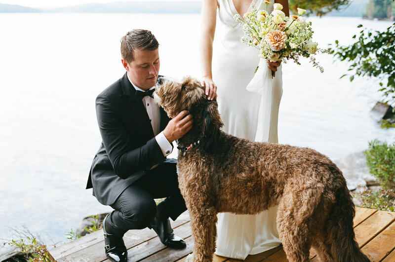 A groom in a black tuxedo pets a large brown dog on a wooden deck by a lake at his Muskoka cottage wedding, while the bride in a white dress holds a bouquet of flowers. Captured beautifully by an Ontario destination wedding photographer, Jennifer van Son, this moment blends love and nature for timeless wedding photography in Toronto.