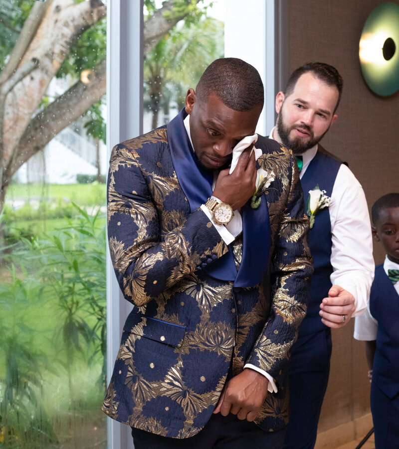 The groom is crying at the wedding while waiting for his bride. The groom is wearing blue and gold with details stitching