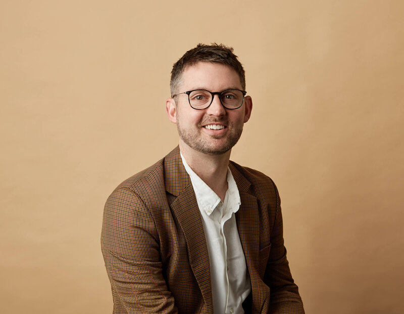 Jessie hands on hips in camel coloured coat against beige background.